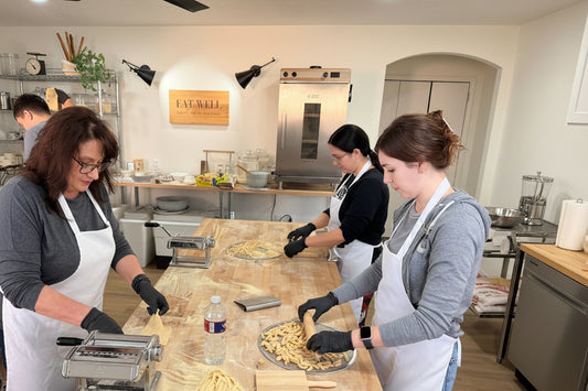 Three people working in a kitchen preparing pasta, with various pasta shapes on the counter.